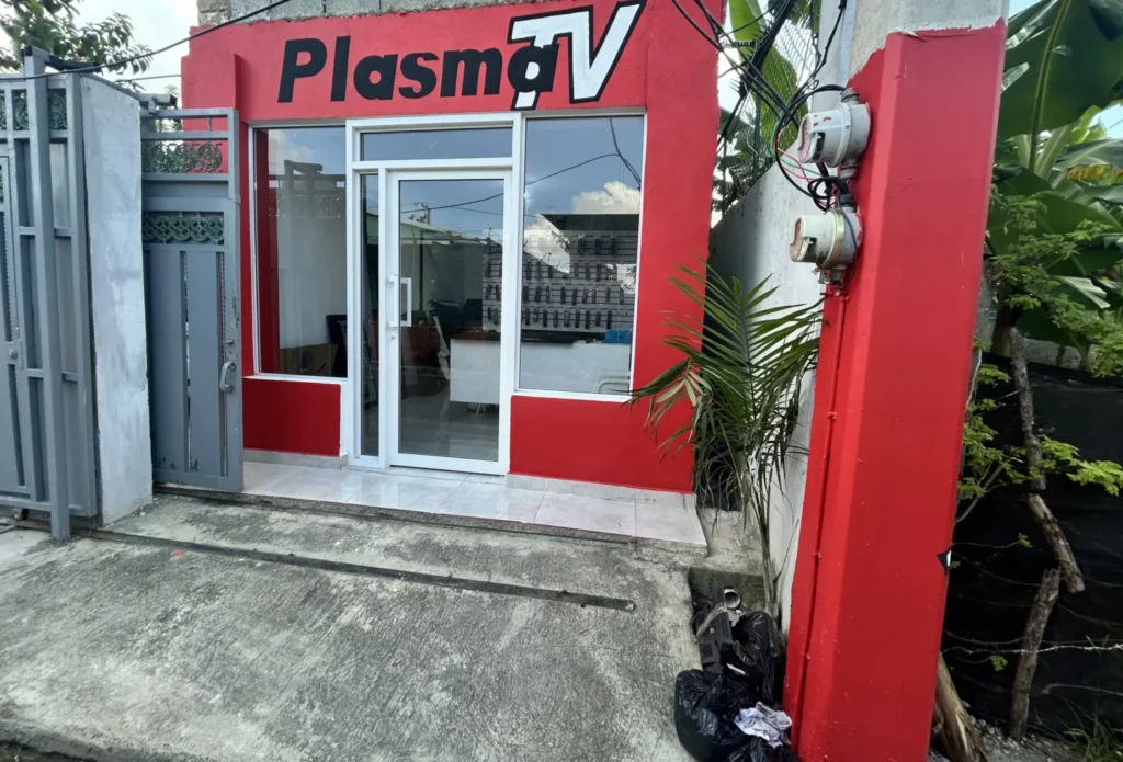 Fachada del local PlasmaTV: edificio con paredes rojas, letrero negro con el nombre en la parte superior, gran ventanal y puerta corrediza de vidrio con marco blanco. En el interior se ven exhibidores de productos y mostradores. A la izquierda hay un portón gris y a la derecha una palmera junto a un poste rojo con medidores eléctricos.